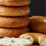 A beautiful stack of freshly baked Montreal bagels, with one sliced open and generously spread with cream cheese on a rustic wooden board.