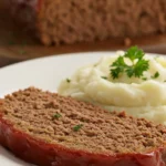 A thick, juicy slice of the finished Lipton Onion Soup Mix Meatloaf recipe on a white plate, showcasing its moist texture and caramelized ketchup glaze, served next to mashed potatoes.