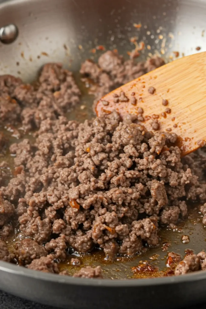 An action shot of ground beef browning in a stainless steel skillet, being broken up with a wooden spatula for the first step of the taco meat recipe.